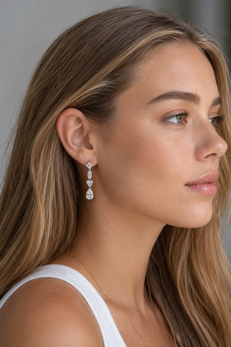 A close-up side profile of a woman with long blonde hair wearing a white gold drop earring with heart-shaped clear stones, paired with a white tank top.